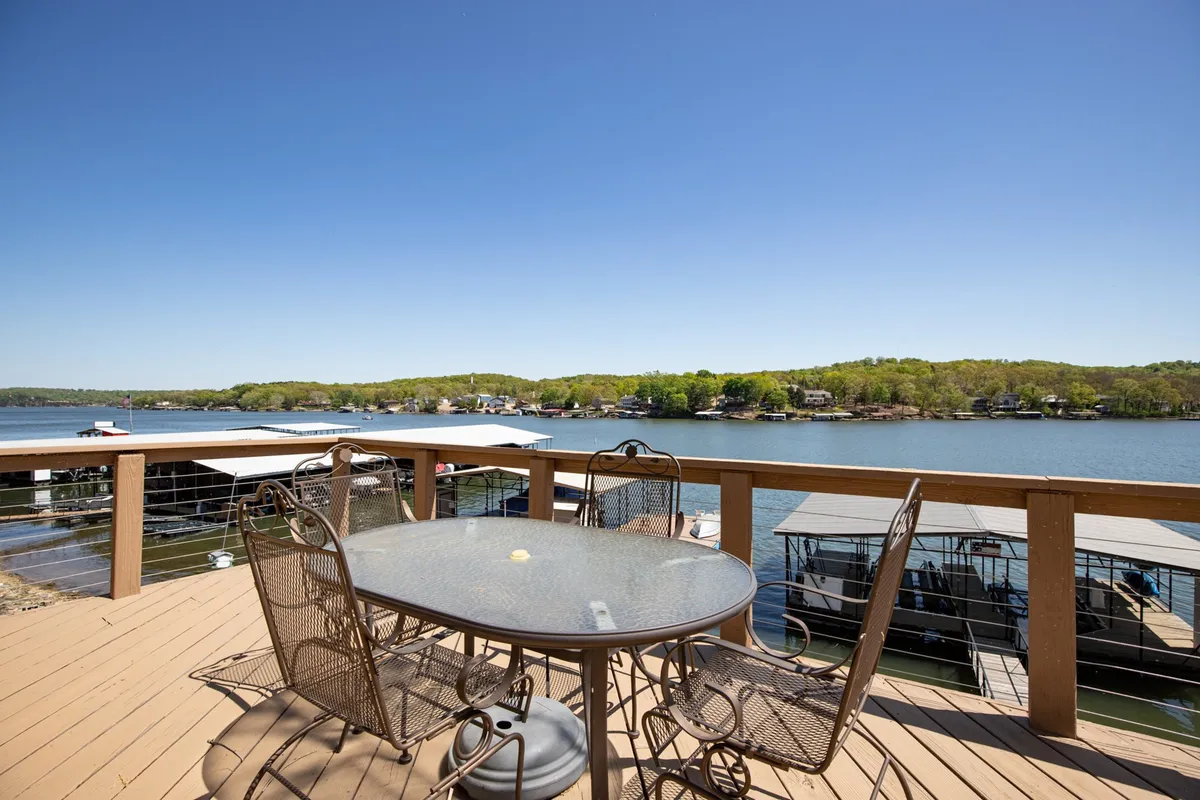 An outdoor dining table with chairs overlooks the lake and boathouses, underlining the joy of al fresco meals with a panoramic waterfront backdrop. The scene captures moments of dining, socializing, and taking in the tranquil views at any time of day.