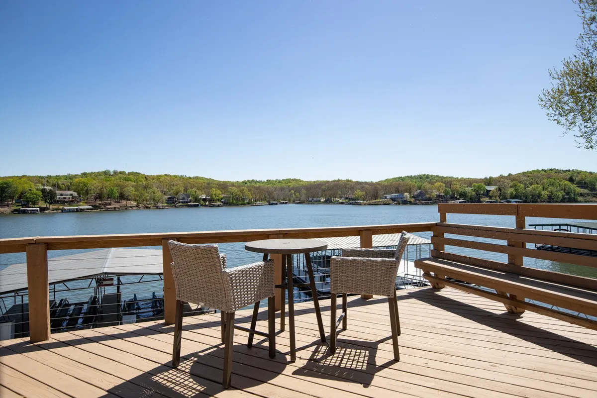 An inviting exterior shot featuring a patio table and chairs overlooking the water, with a backdrop of distant houses and trees. The scene feels serene and social, perfect for outdoor dining or gathering with friends and family.