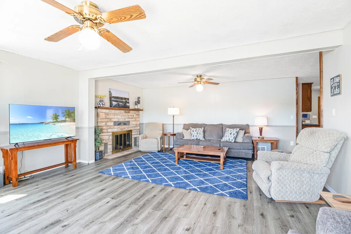 A welcoming living room with a comfortable gray sofa, blue geometric rug, wooden coffee table, large flat-screen TV, and a classic brick fireplace. Large windows offer views outside, and the space feels ideal for both relaxation and productivity, with smart tech and cozy touches.