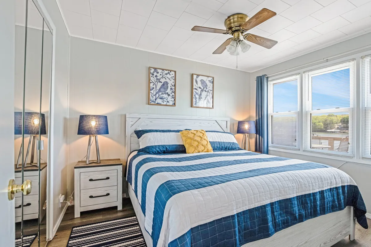 A bright, peaceful bedroom featuring a bed with blue striped bedding, decorative pillows, nightstands with lamps, framed artwork on the walls, and large windows with curtains that allow sunlight to stream in.