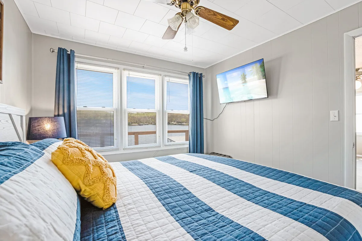 A comfortable bedroom with a bed adorned with a striped quilt, large windows offering ample natural light, and a wall-mounted television for cozy evenings. The space feels calm and inviting.