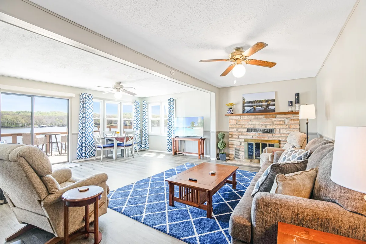 This image features a welcoming open-plan living area with a comfortable seating area, a stone fireplace, large windows overlooking the lake, and a dining space. The space is styled for relaxation and socializing with ample natural light and panoramic lake views visible through the windows.