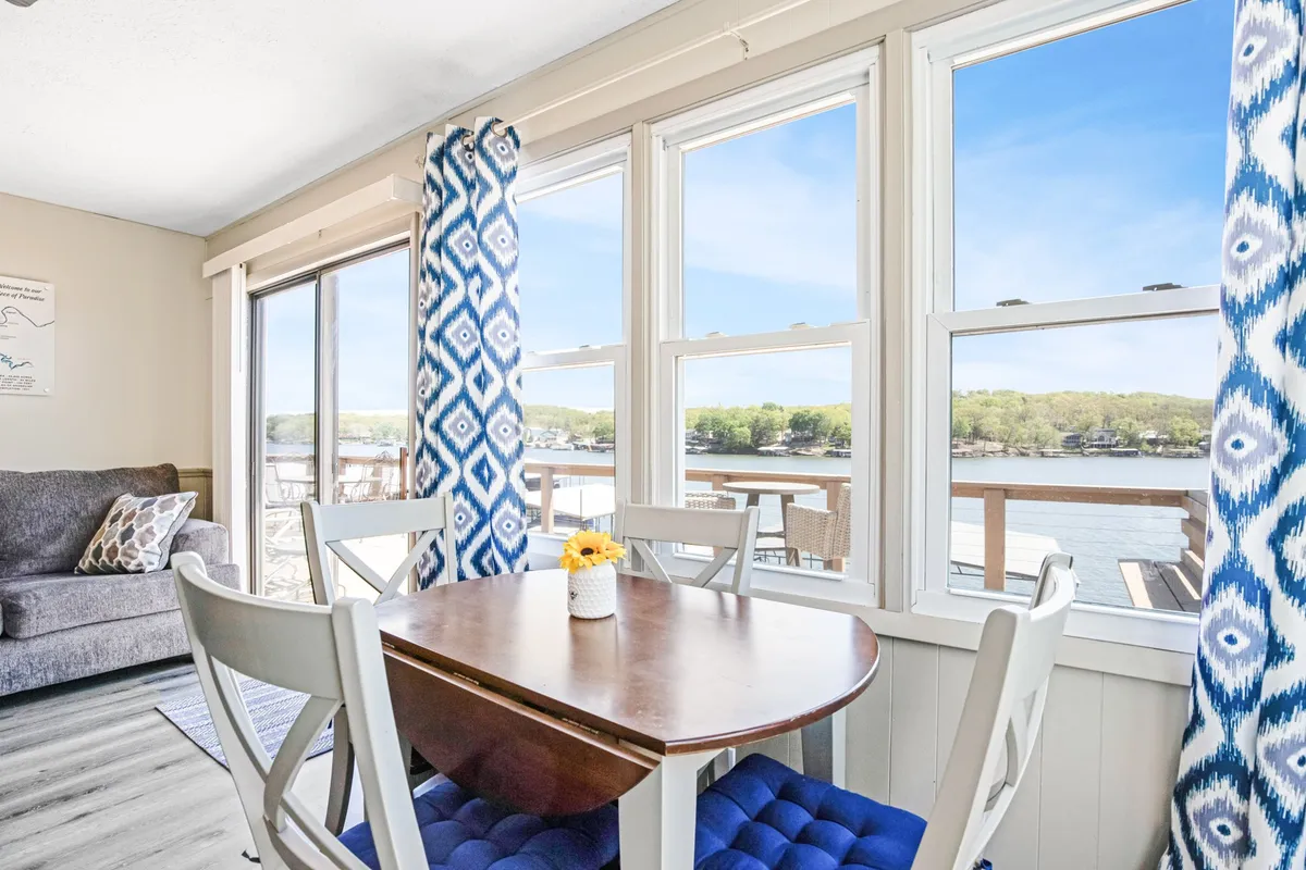 A cheerful dining area set beside large windows, with a dining table ready for family meals or board games, a cozy sofa, and decorative curtains. The scene captures the light-filled social hub of the home, inviting guests to savor meals and moments together while admiring the water just outside.