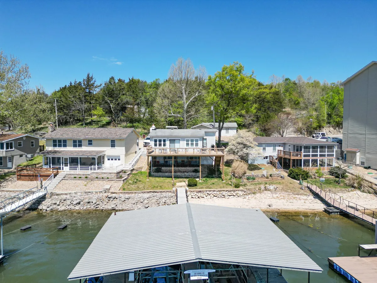 A tranquil view of houses along the shoreline with a prominent dock extending over the calm lake waters. Lush green trees frame the scene, and the overall setting radiates peacefulness. The vantage point hints at opportunities for morning coffee on the deck, evening gatherings, and quiet reflection by the water.